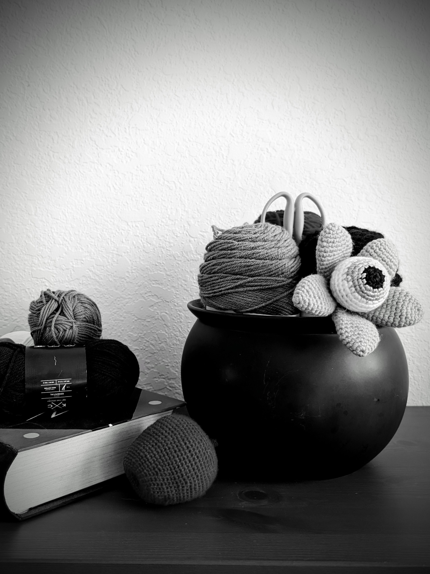 A black cauldron filled with yarn balls and crochet pieces beside a book, in moody black and white